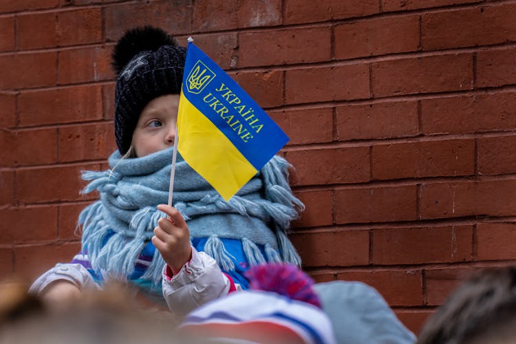 Child In Black Knit Hat And White Scarf Leaning Against A Brick Wall Holding A Flag