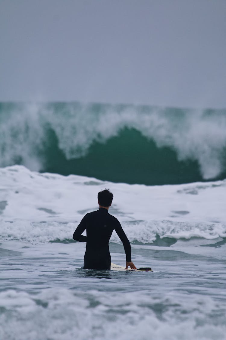 Man Standing In Sea Holding Surfboard Looking At Big Wave 