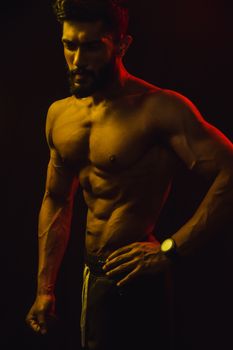 Strong and muscular man posing in dimly lit studio with dramatic lighting, showcasing fitness and strength.