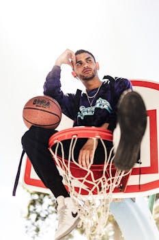 Confident man sitting on a basketball hoop outdoors, holding a ball, with ample copy space around.