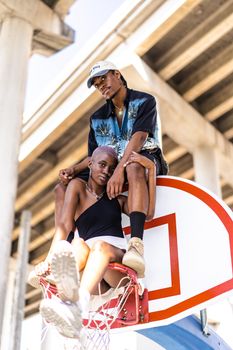 Young couple playfully sitting on a basketball hoop under a bridge, showcasing urban lifestyle.