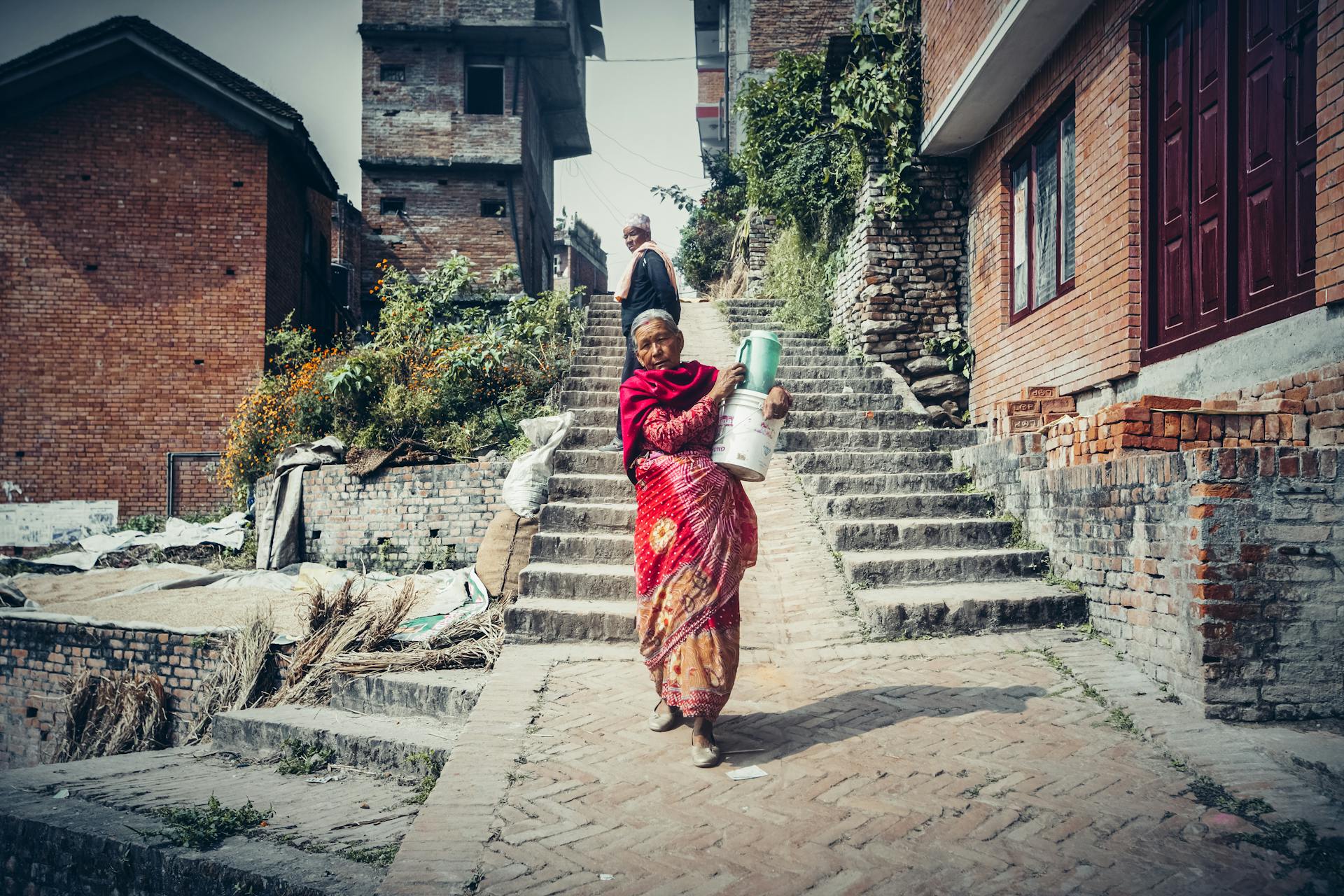 Elderly woman in traditional clothing carries a gas cylinder up brick steps in a rural town.