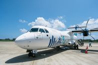 Manta Air turboprop plane parked on airport runway under clear blue skies with distant passengers.