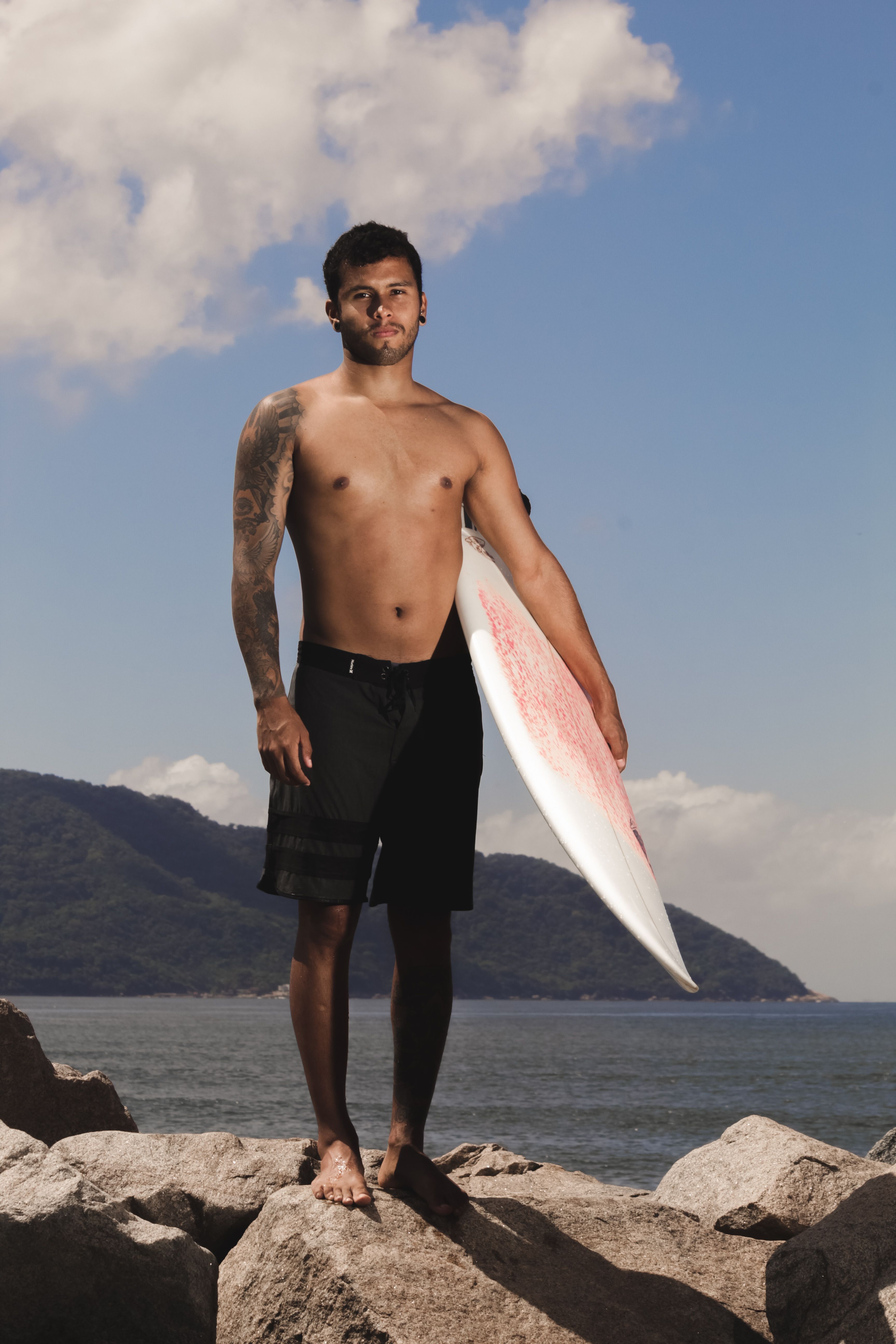 Man standing shirtless with surfboard on rocky beach against blue sky backdrop.