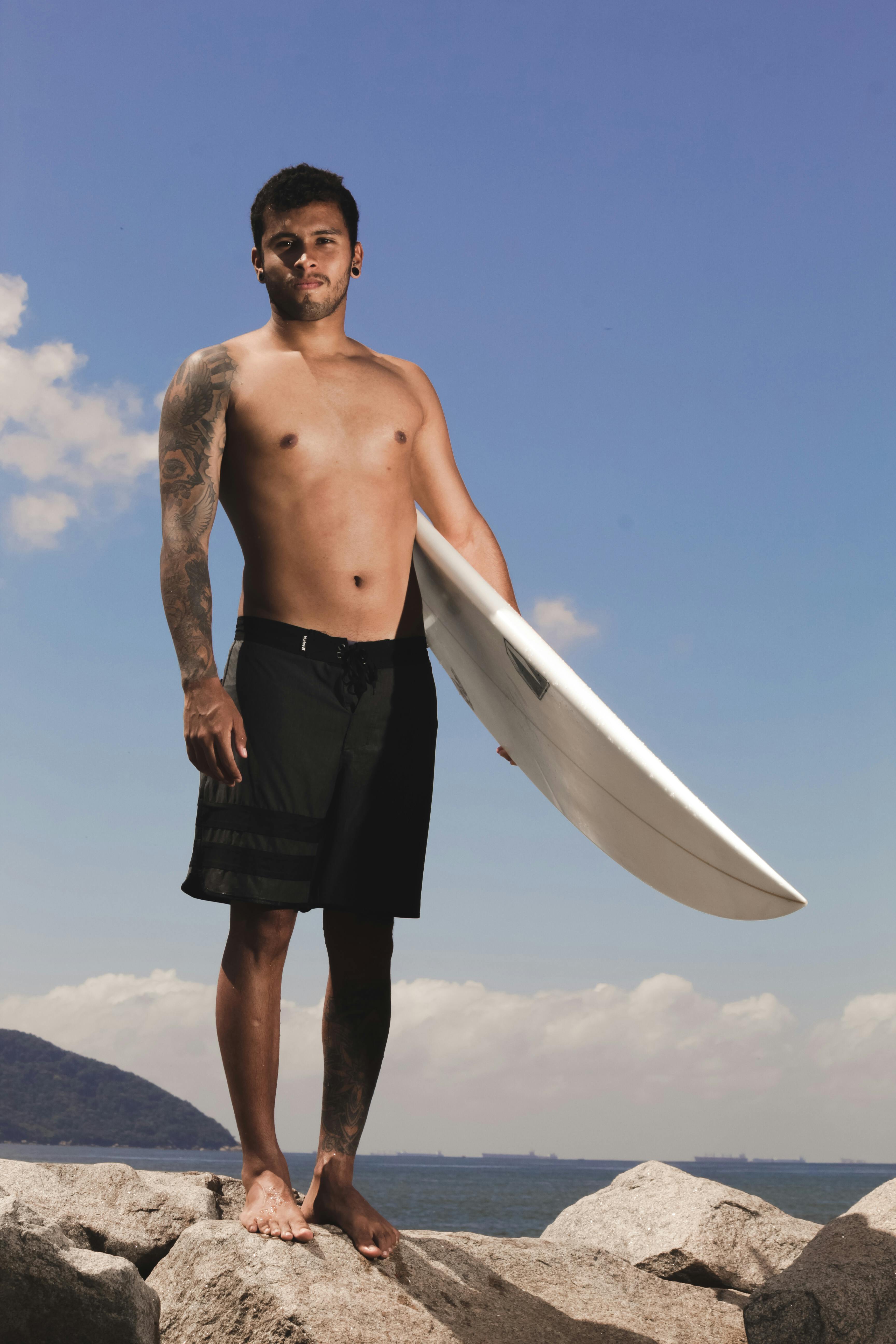 Tattooed surfer holding a surfboard on rocky beachfront under a clear sky.