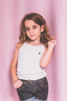 Young girl in denim shorts and white shirt posing confidently against a pink background.