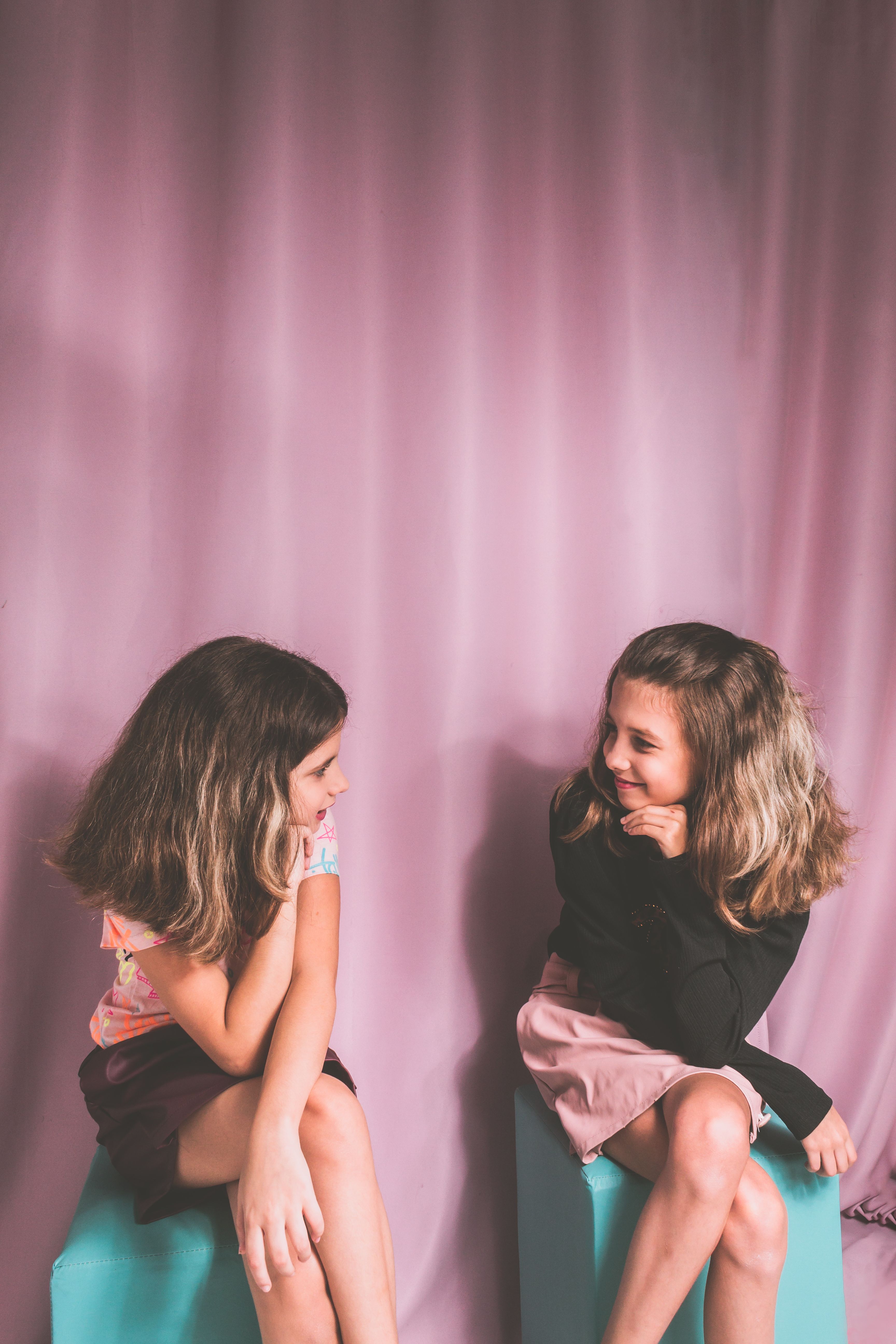 Two young girls sitting and conversing in a studio with a soft pink background.