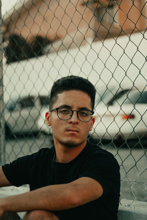 Free Portrait of a man with glasses sitting in front of a chain-link fence and parked cars. Stock Photo