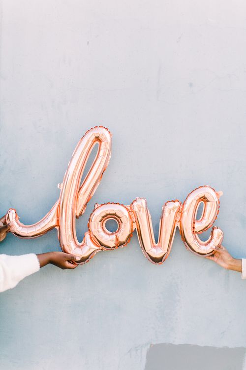 Free Hands hold a rose gold balloon spelling 'love' against a blue wall, perfect for romantic themes. Stock Photo