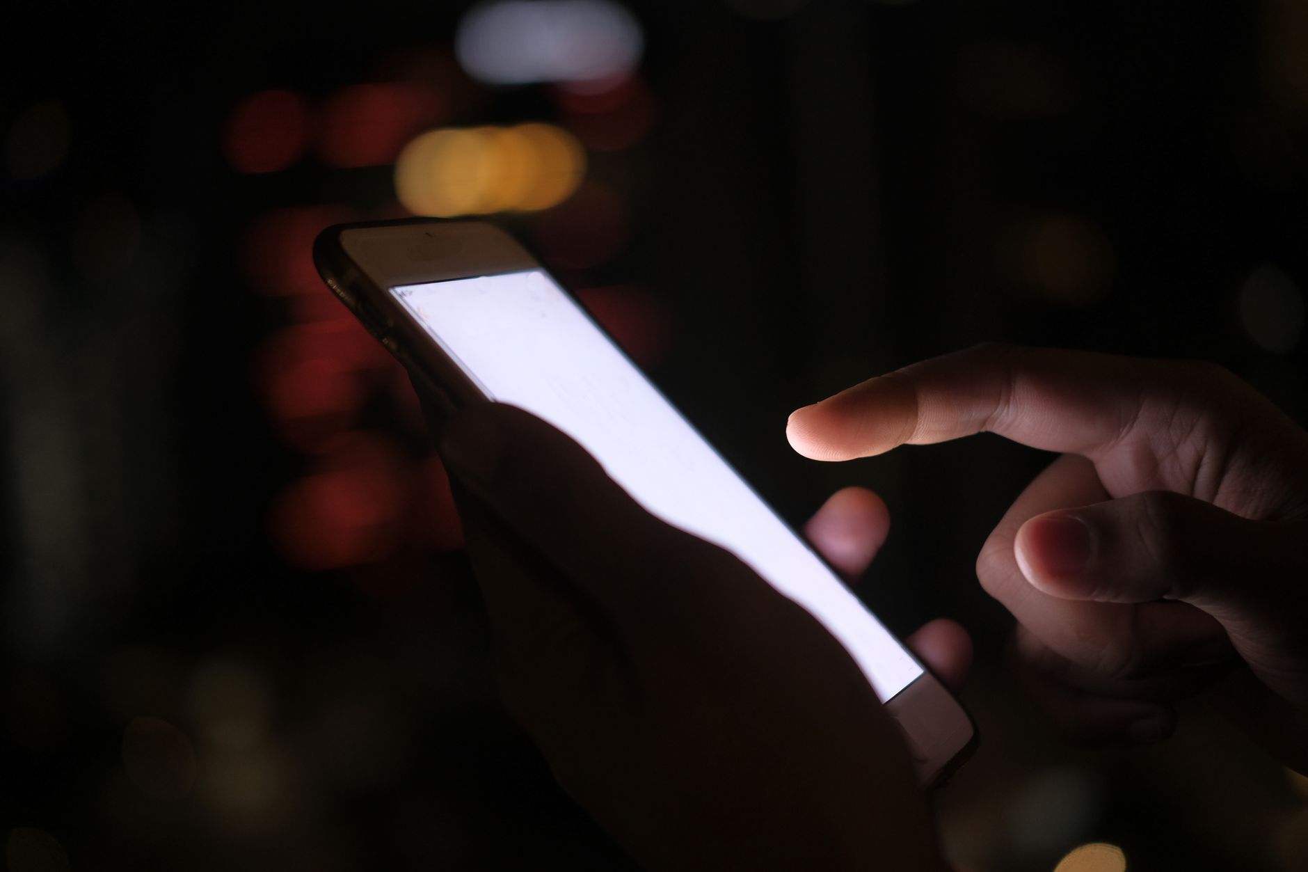 Close-up of a hand using a smartphone in the dark with blurred city lights in the background.