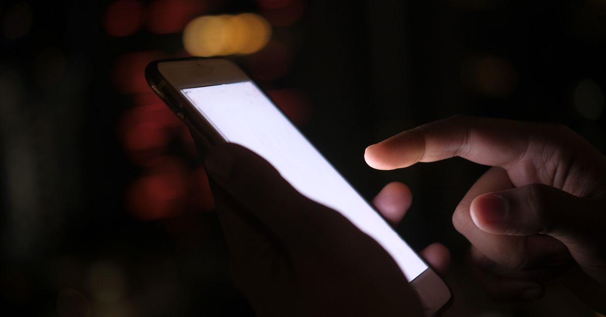 Close-up de uma mão usando um smartphone no escuro com luzes da cidade desfocadas ao fundo.
