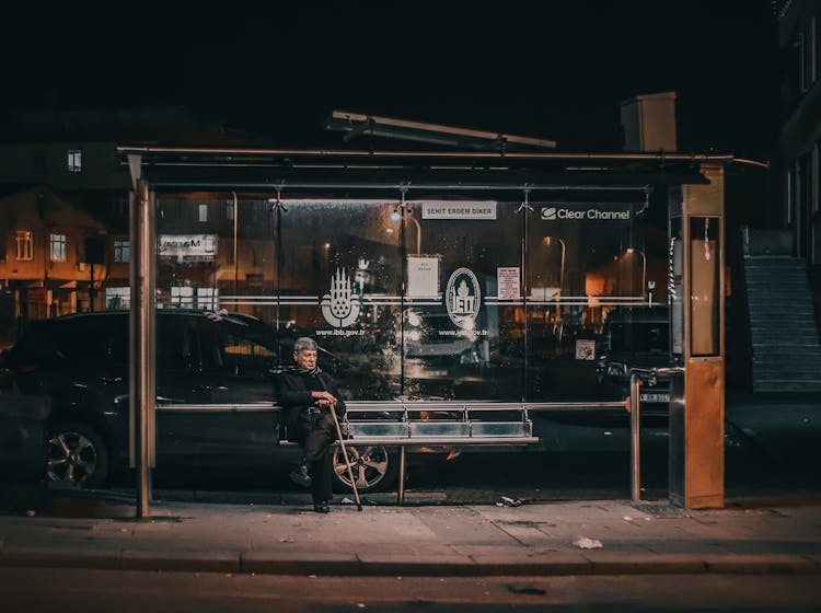 An Elderly Man Sitting On A Bus Stop At Night