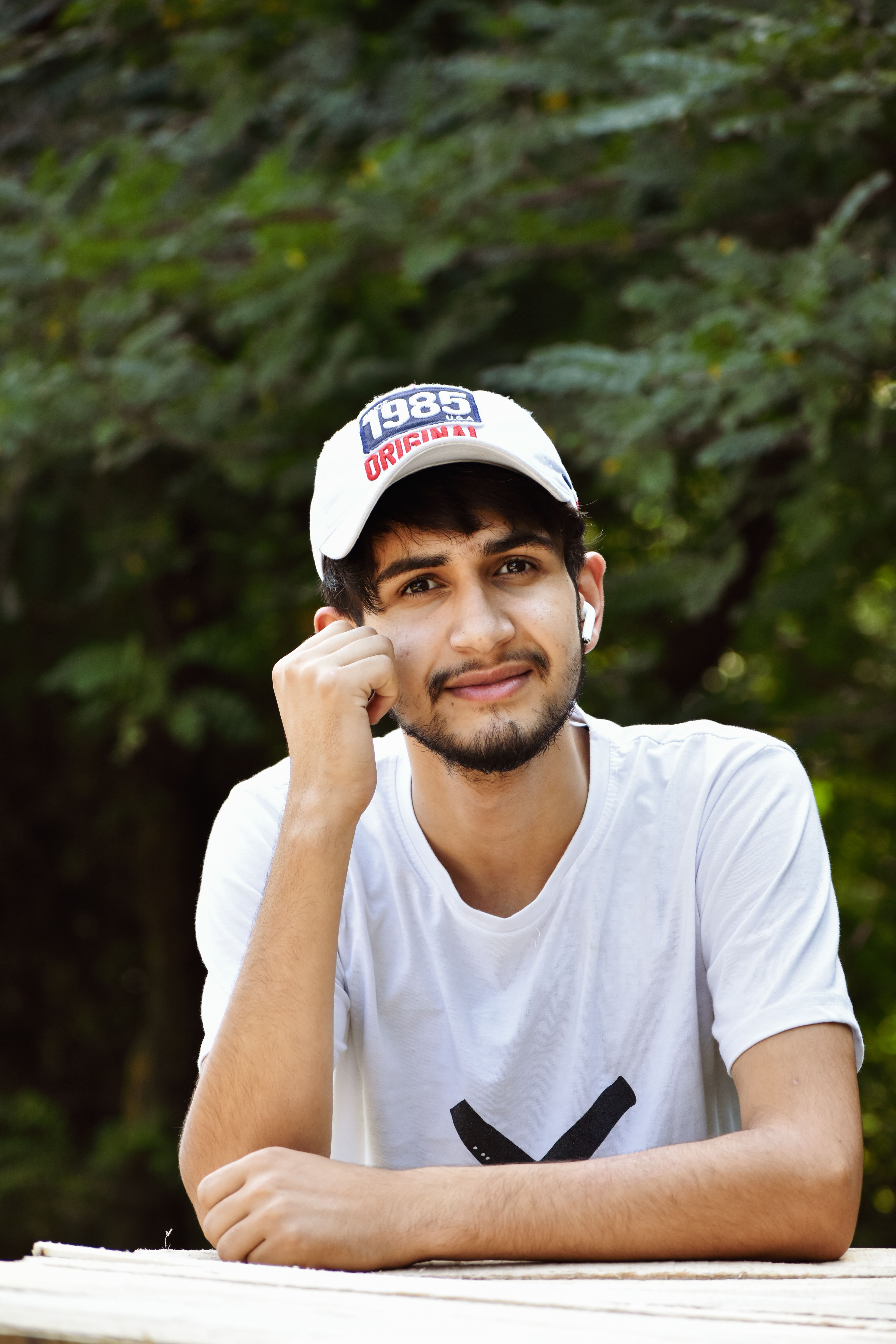Young man in casual attire smiling outdoors with a white cap.