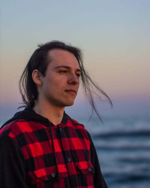Free A thoughtful young man in a checkered jacket gazes into the distance by the ocean at sunset. Stock Photo