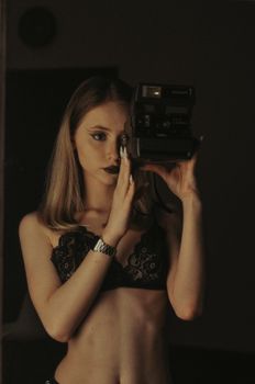A woman in lingerie poses with a vintage Polaroid camera for a moody self-portrait indoors.