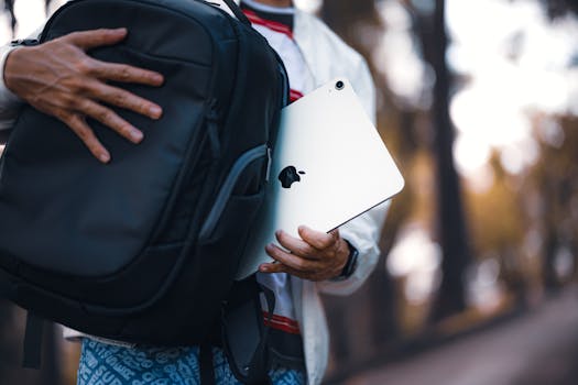Person holding a tablet near a backpack with a blurred outdoor background, highlighting technology and portability.