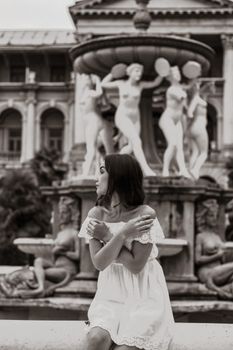 A woman in a white dress poses elegantly near a classical fountain in a black and white photograph.