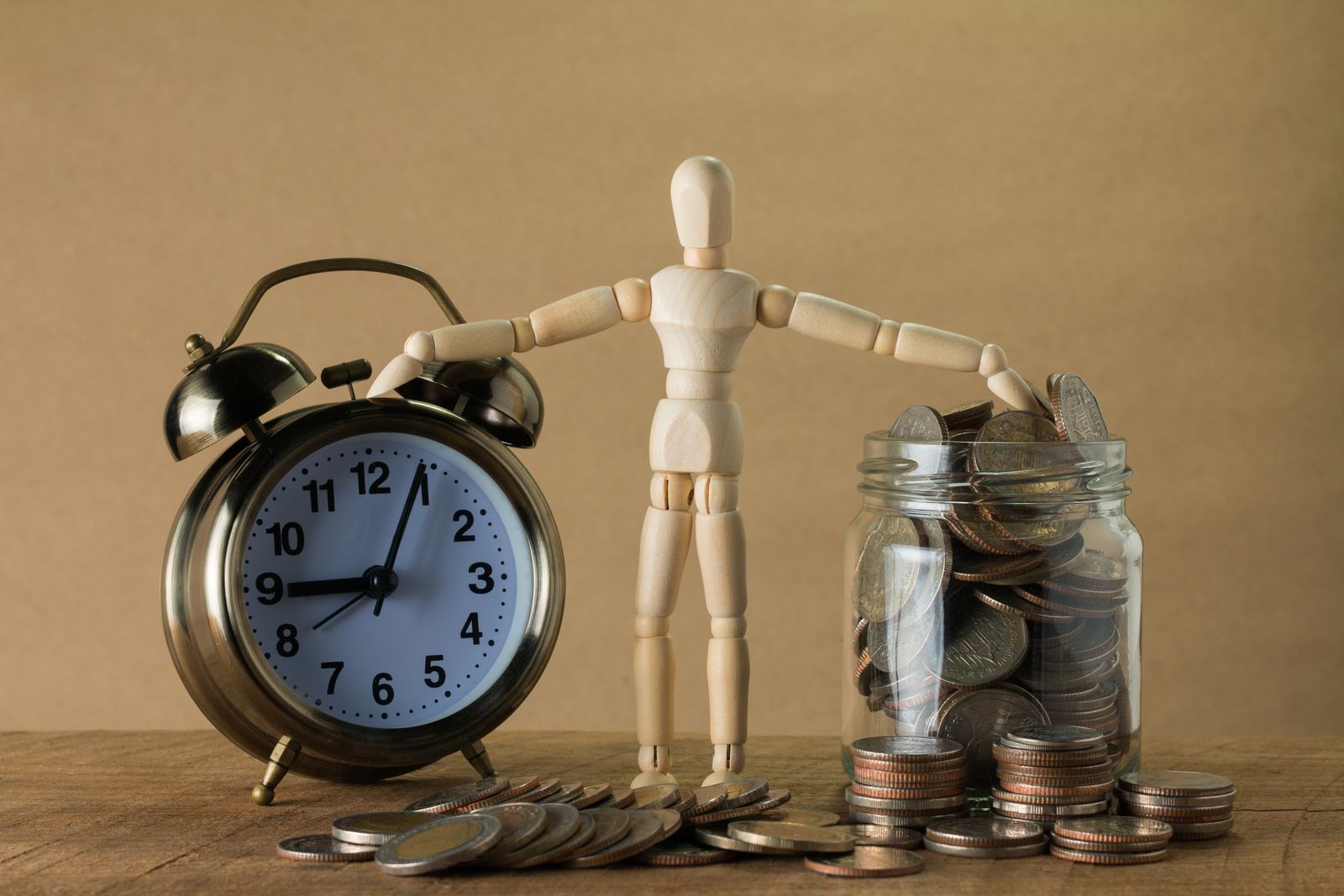 Free Wooden mannequin holding an alarm clock and a jar full of coins, symbolizing time and money. Stock Photo
