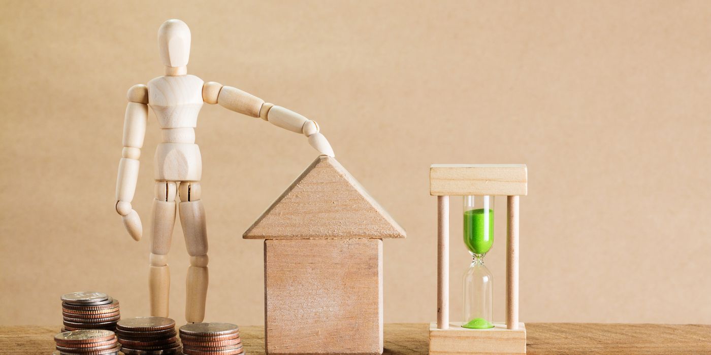 Wooden mannequin with house model, coins, and hourglass symbolizing financial planning.