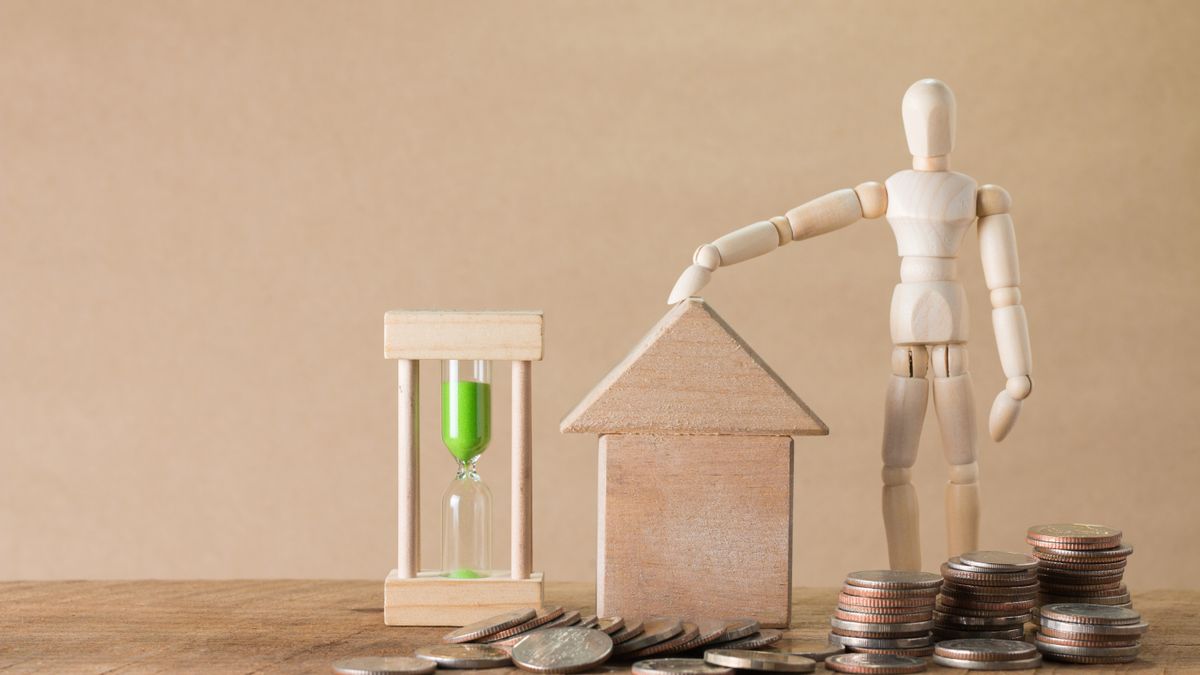 Wooden mannequin with house model, coins, and hourglass on a wooden table, symbolizing time and financial growth.