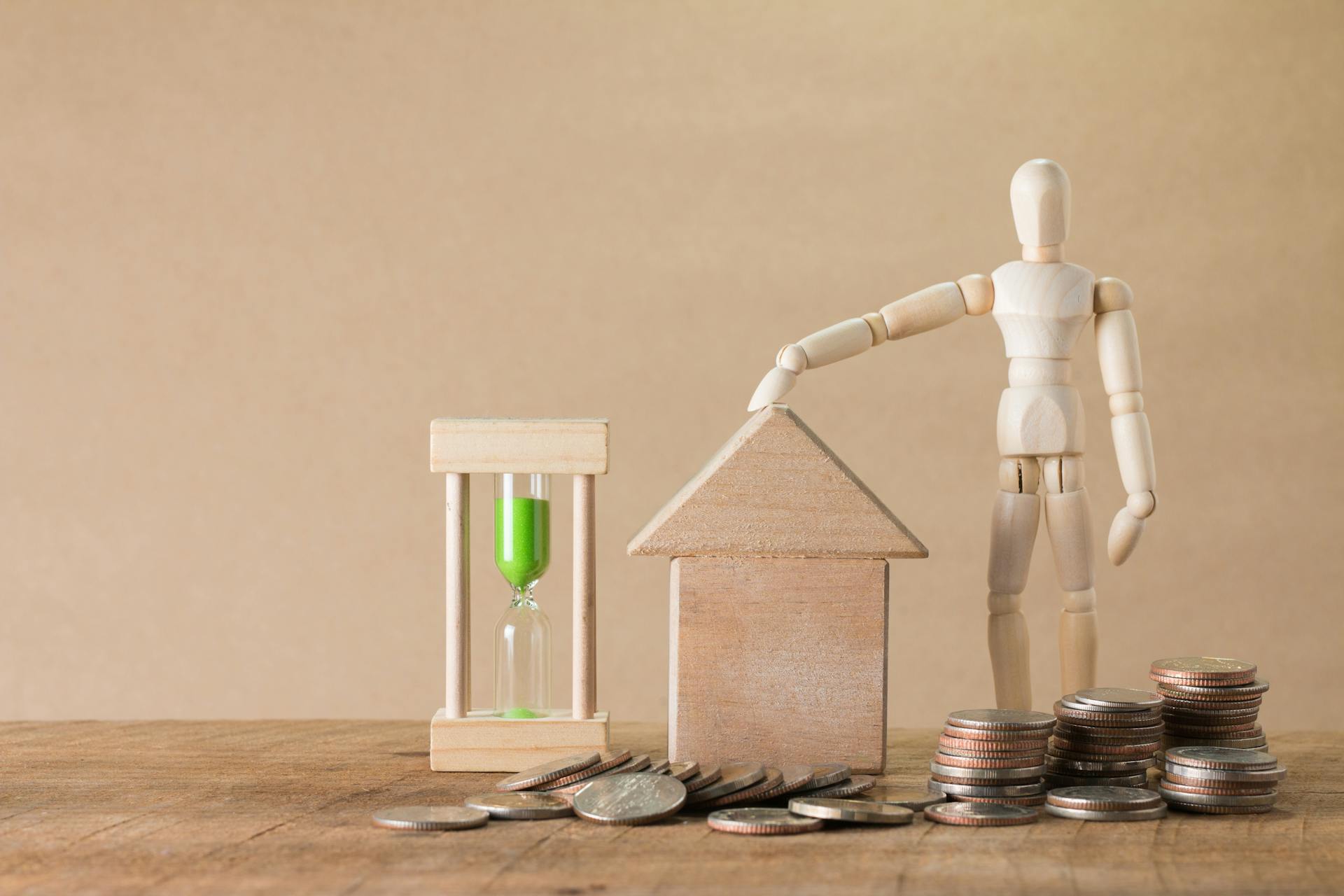 Wooden mannequin with house model, coins, and hourglass on a wooden table, symbolizing time and financial growth.