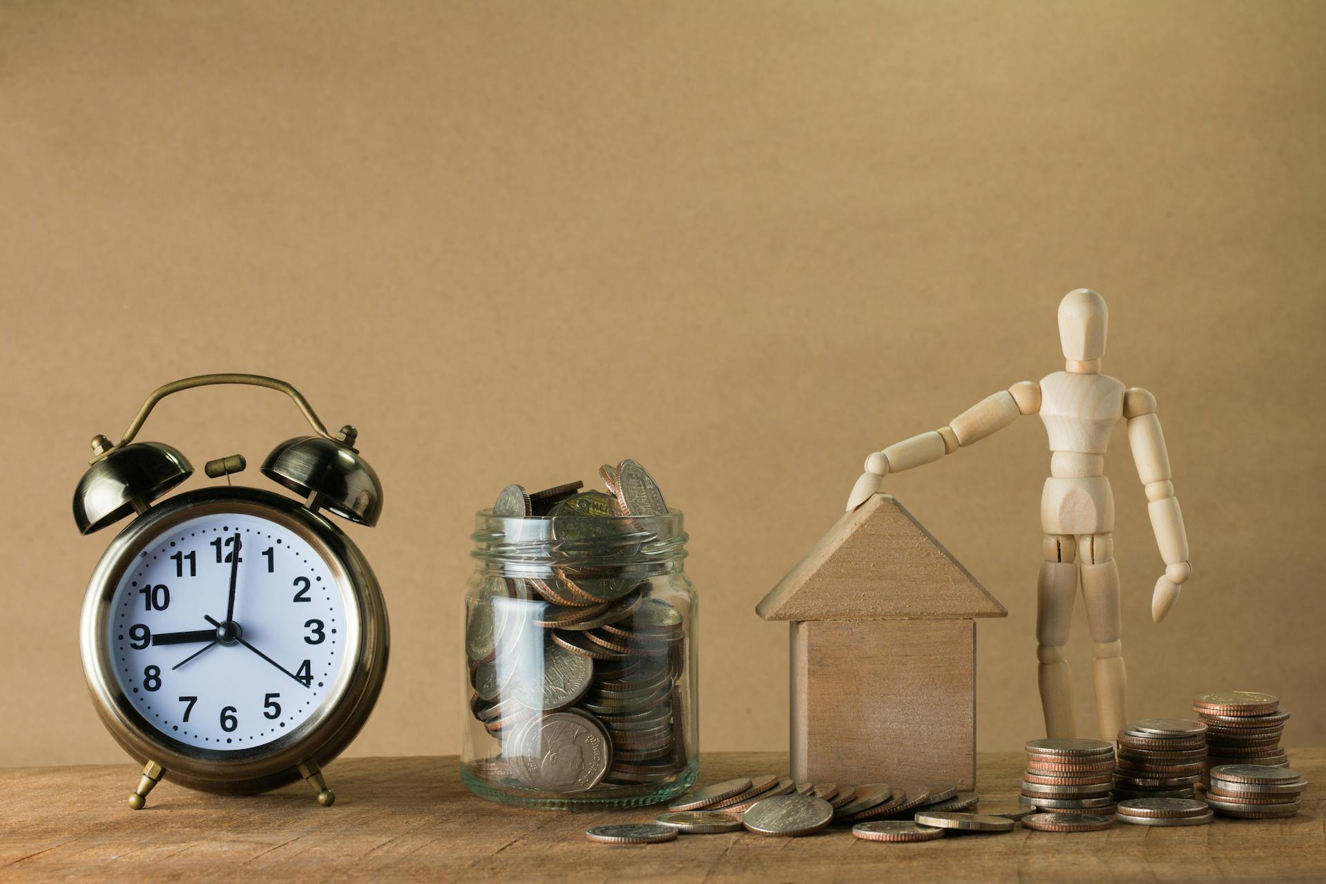 Wooden mannequin with a house, coins, and clock symbolizing time and financial planning.