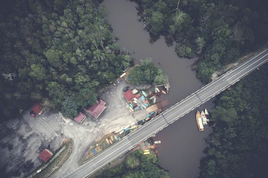 从空中俯瞰，一座桥梁横跨森林覆盖的河流，可见船只与道路基础设施