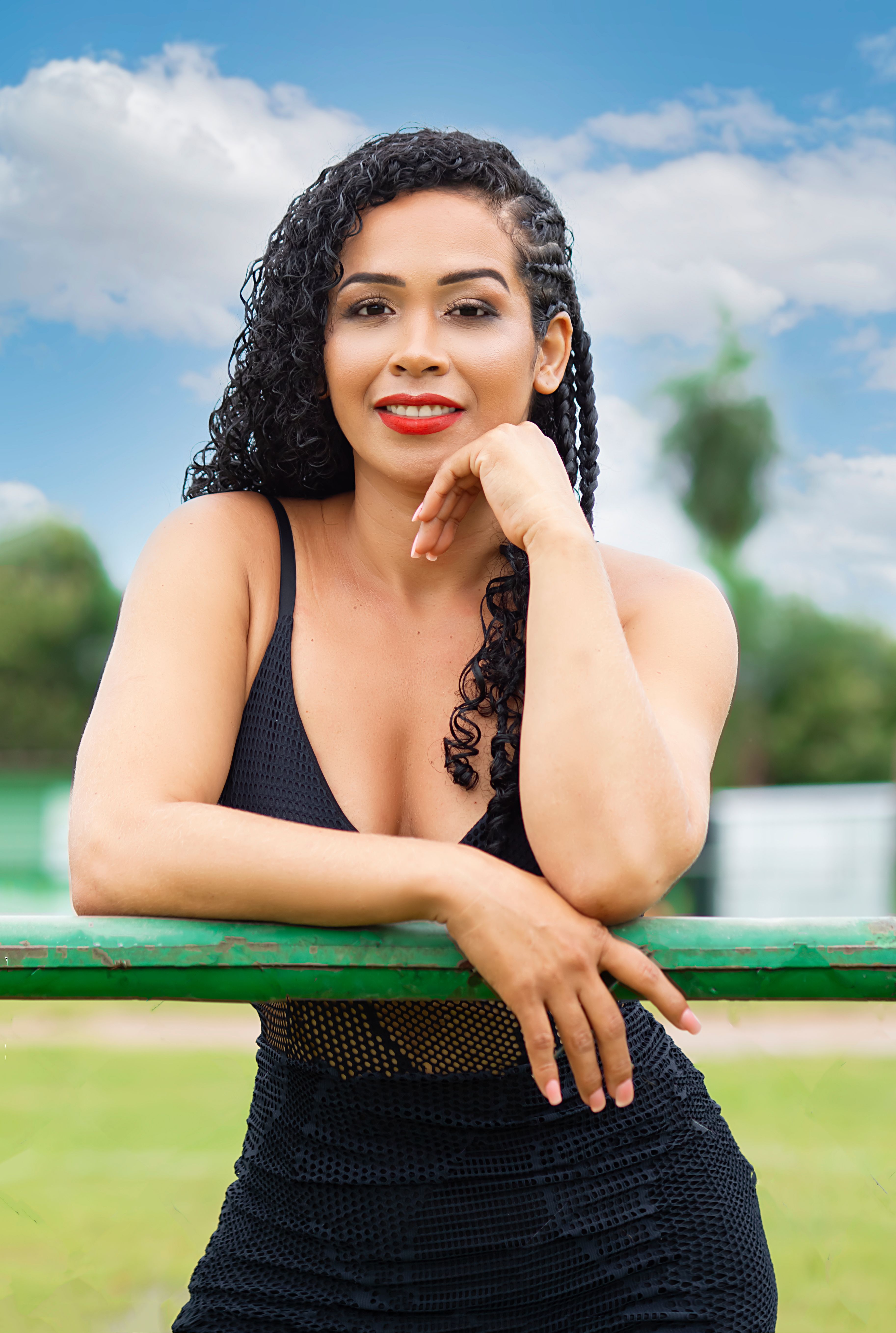 A confident woman with curly hair poses outdoors in a black dress under a blue sky.