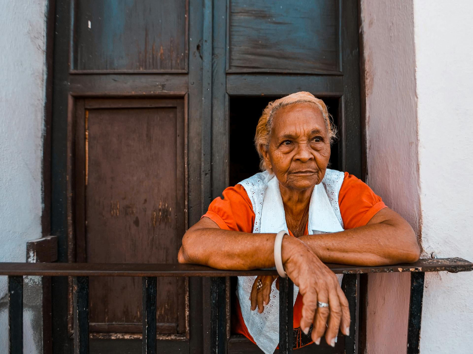 Senior woman in orange shirt leaning on a metal railing balcony.