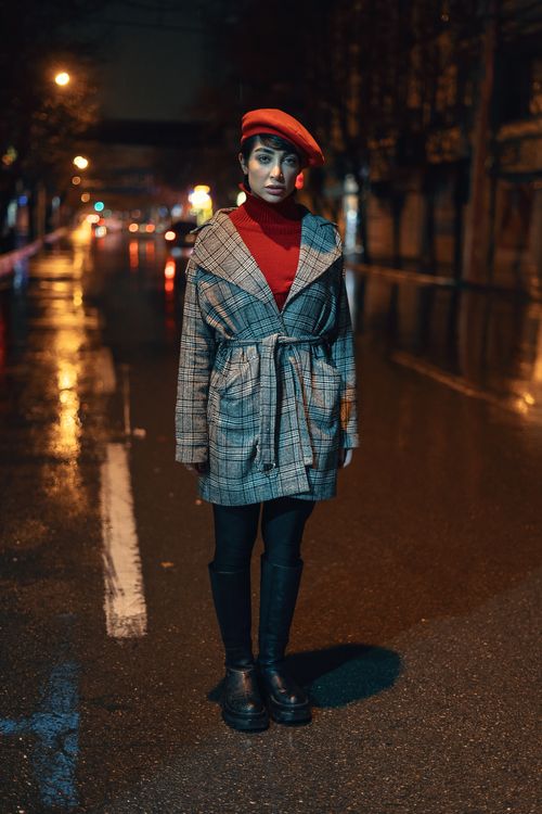 Free A fashionable woman in a plaid coat and red beret stands on a wet city street at night. Stock Photo