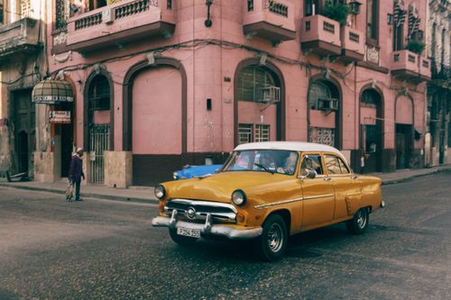 Free Classic yellow car passes by a historic building on a street in Havana, Cuba. Stock Photo