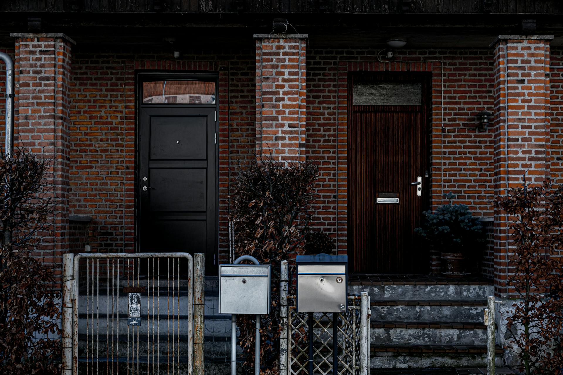 A brick building facade with two distinct doorways and metal fencing, featuring mailboxes and urban elements.
