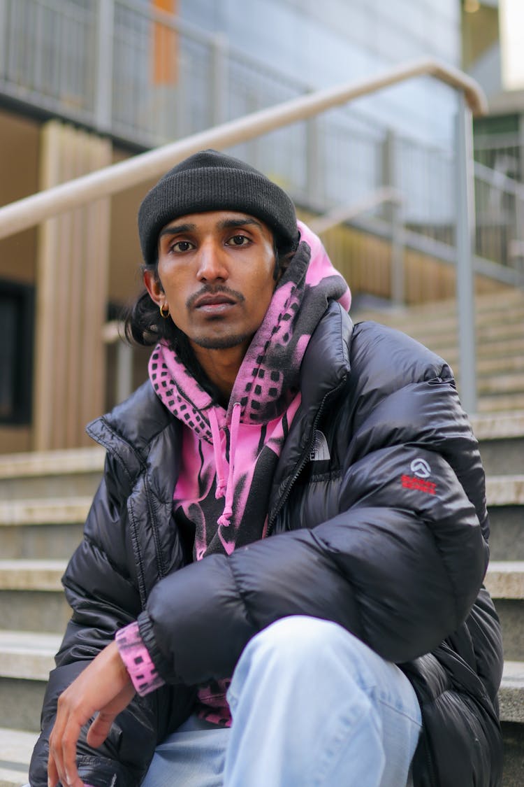 Young Male Model Sitting On Stairs In Beanie And Pink Hoodie And Black Jacket