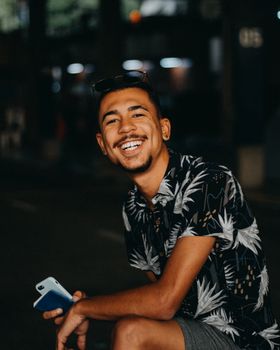 Portrait of a young man smiling with a smartphone in hand, on a dimly lit city street at night.