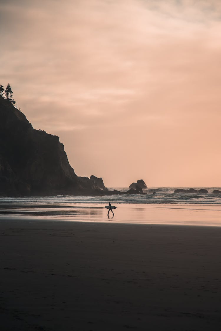 A Man Walking On The Beach Carrying A Surfboard