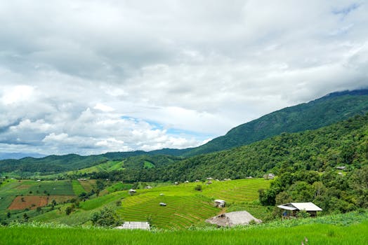 A picturesque landscape of lush green fields, hills, and farmhouses under a cloudy sky.
