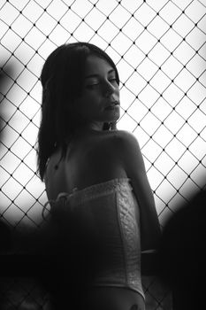 Elegant black and white portrait of a young woman wearing a corset, posing by a chain-link fence.