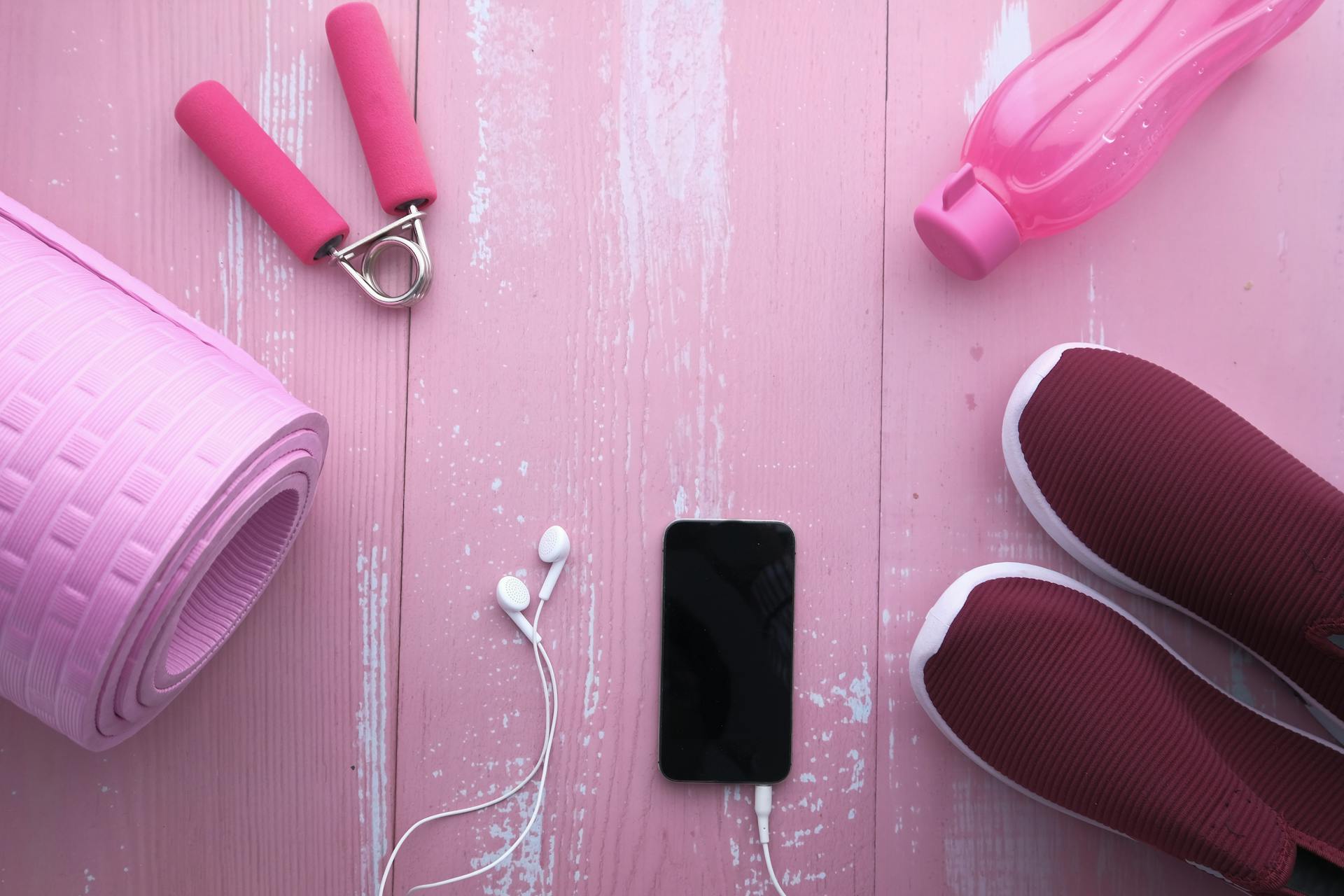 Top view of fitness accessories including yoga mat, shoes, water bottle, and smartphone on pink wood.