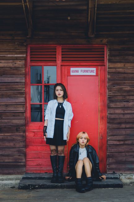 Two fashionable women stand and sit by a rustic red wooden door labeled Kaina Furniture.