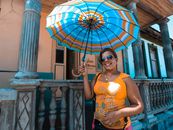 A cheerful woman poses with a colorful umbrella in front of a traditional building on a sunny day.