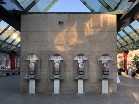 Four coin operated payphones in urban area of Milan, Italy, reflecting sunlight.