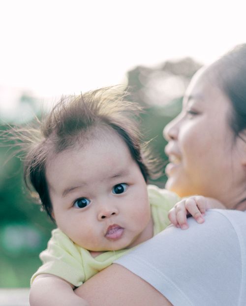 Free A cute baby with expressive eyes being held by a smiling mother in a natural outdoor setting. Stock Photo