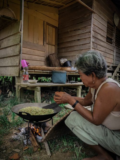 Free An elderly woman cooks over a fire outside a wooden house, creating a rustic and homey atmosphere. Stock Photo