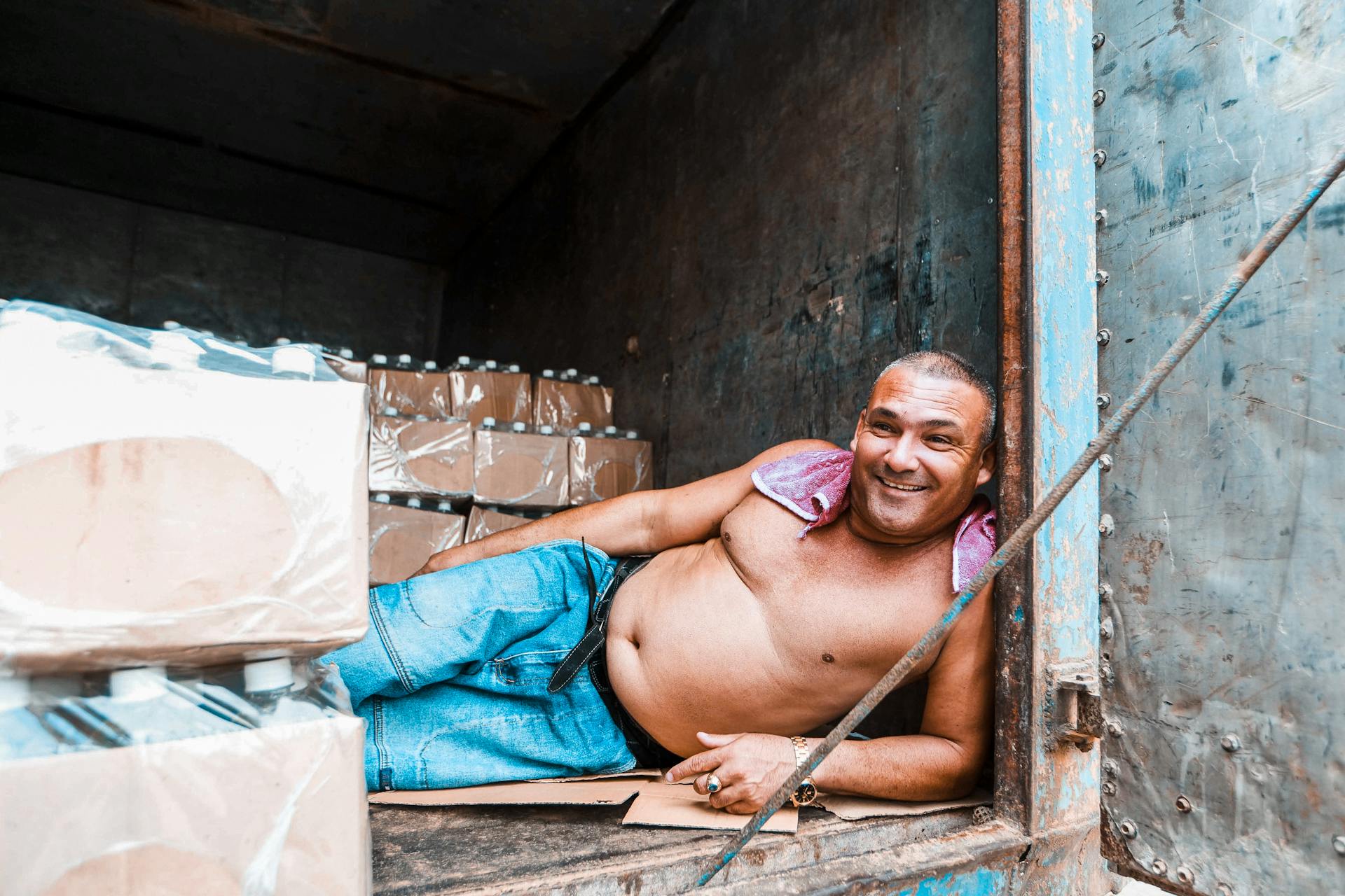 Smiling man relaxing on boxes inside a cargo container, wearing casual attire.