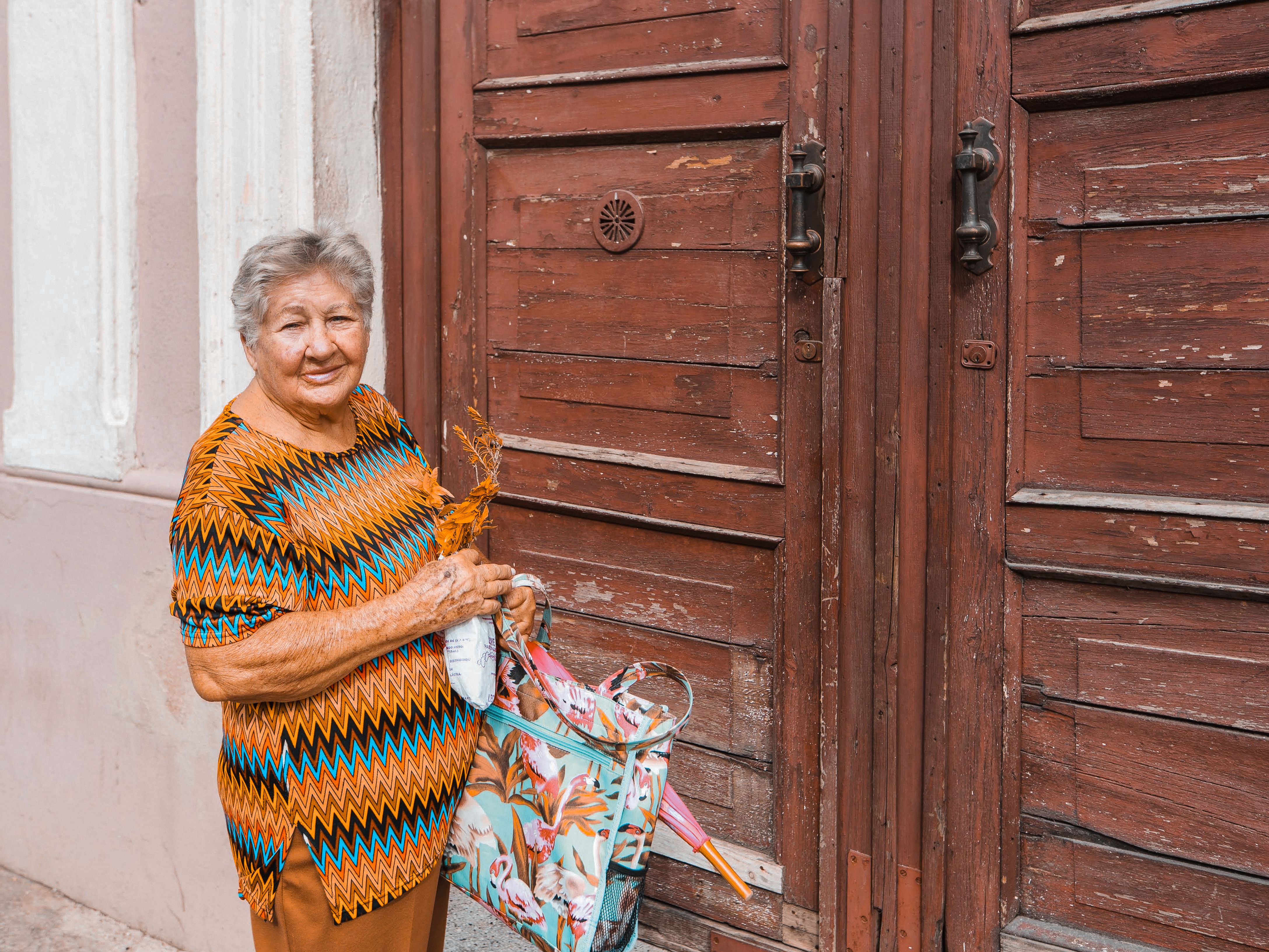 Elderly woman standing by a vintage wooden door with a colorful bag, exuding warmth