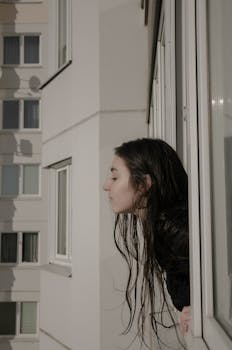 A woman with dark hair leaning out of a window in an urban apartment building.