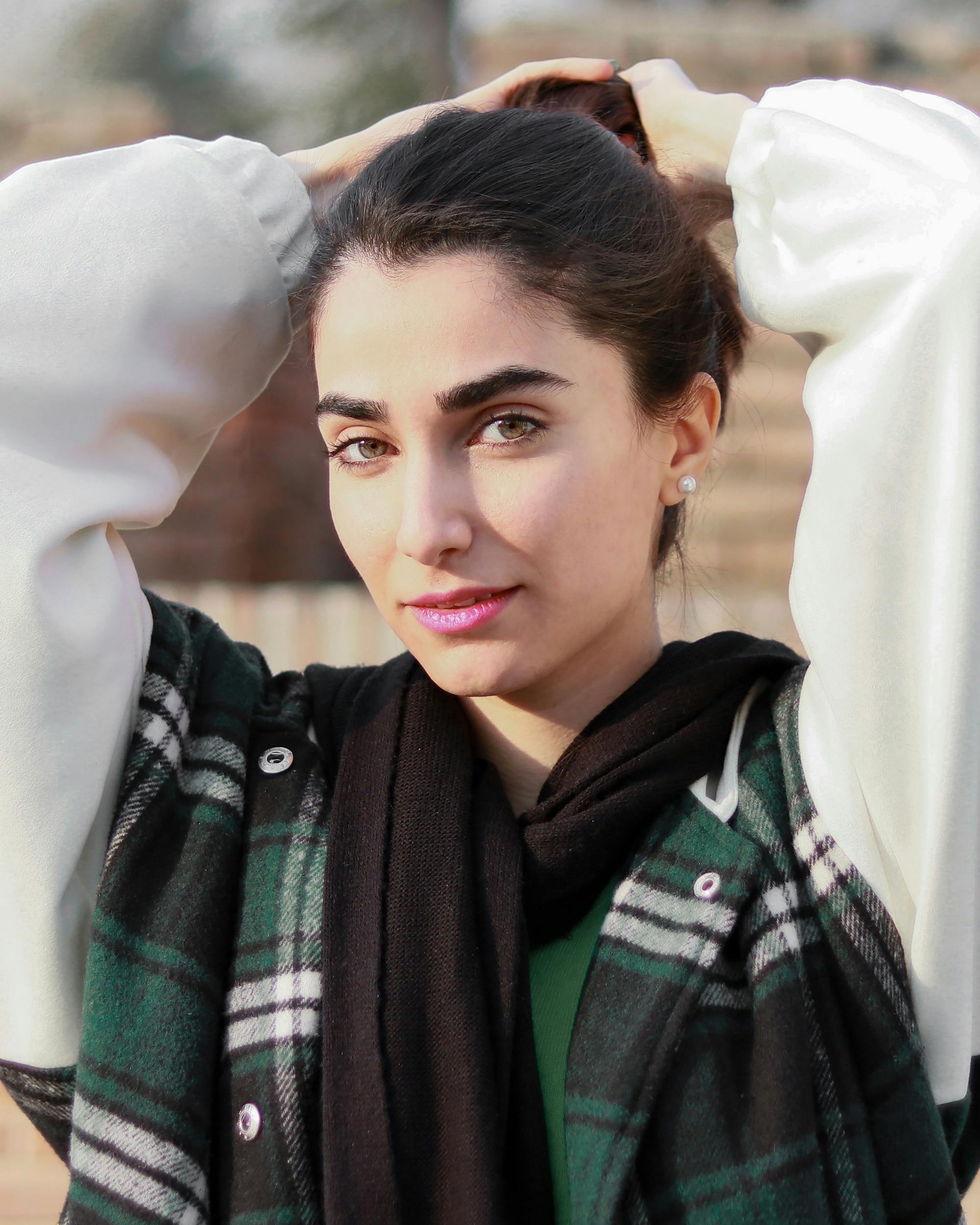 Close-up portrait of a young woman with hands on head, wearing a plaid coat outdoors.