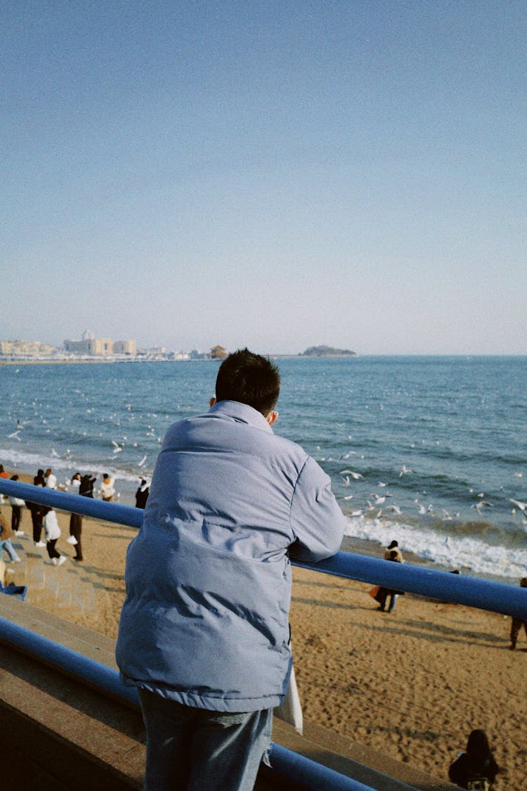 Back View Of Man Leaning On A Barrier And Looking At The Sea 