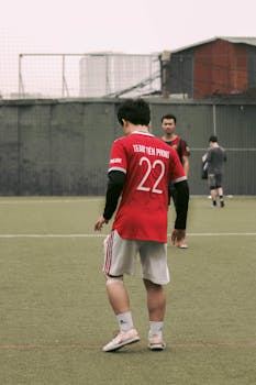 Back view of a soccer player in a red jersey during a game on an urban field.