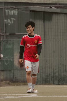 Asian male adult in a red soccer jersey walking on an outdoor field.